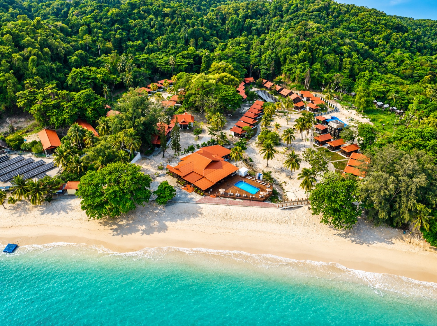 Aerial view of The Heritage Kapas Island Resort: orange-roofed timber villas among palms, a pool, and white sand meeting turquoise water.