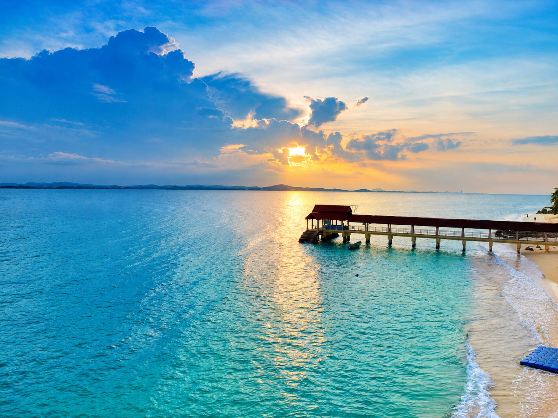 Sunset over the turquoise water at Pulau Kapas, with a wooden jetty reaching out from the beach.