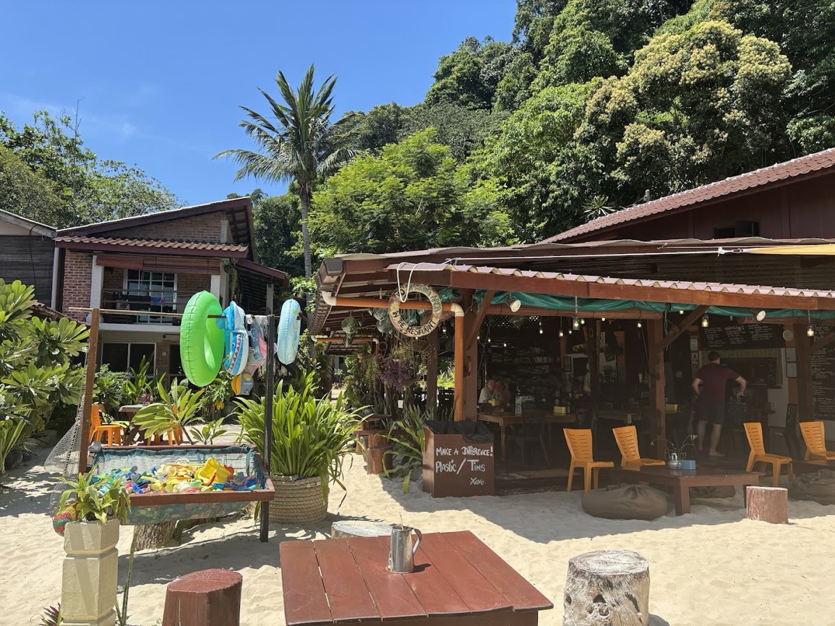 The KBC beach restaurant in daylight: an open-sided timber pavilion with chalkboard menus, wooden chairs on the sand and swim rings hanging from a frame.