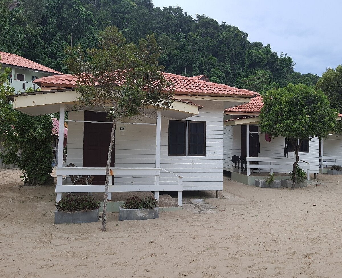 White timber chalets with terracotta roofs at Kapas Coral Beach Resort, standing on the sand below a jungled hillside.