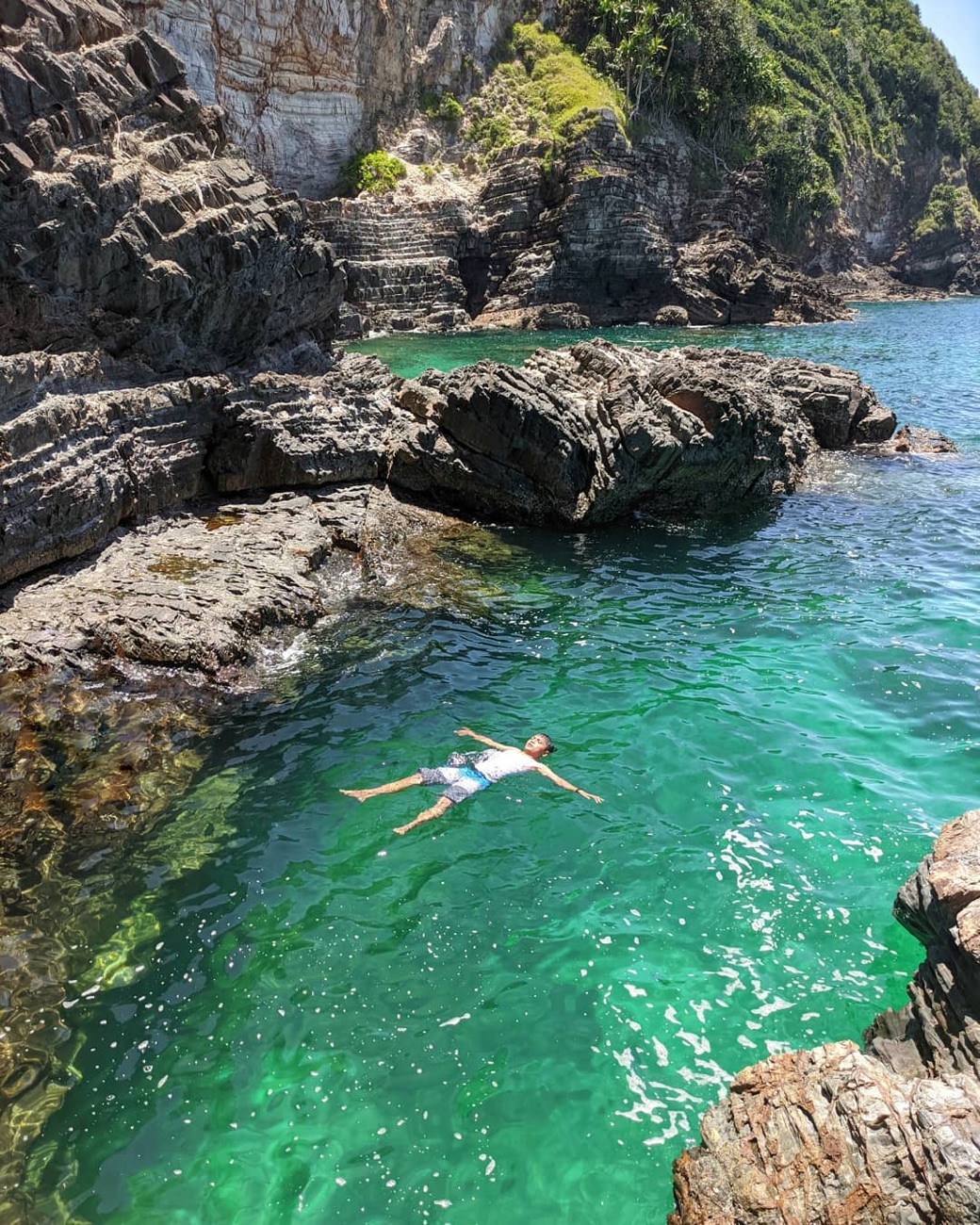 A swimmer floating on their back in clear green water inside a rocky cove on Pulau Kapas, with layered rock ledges rising around them.