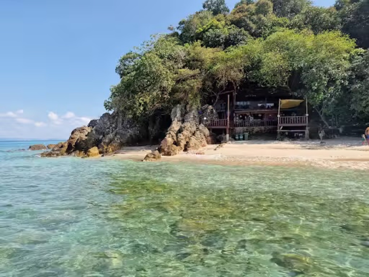 The secluded cove at Kapas Turtle Valley: clear shallow water over rock, a rocky headland, and a small timber building above the sand with a kayak beside it.